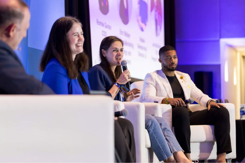 Four panelists seated on stage, one woman speaking into a microphone at a conference.